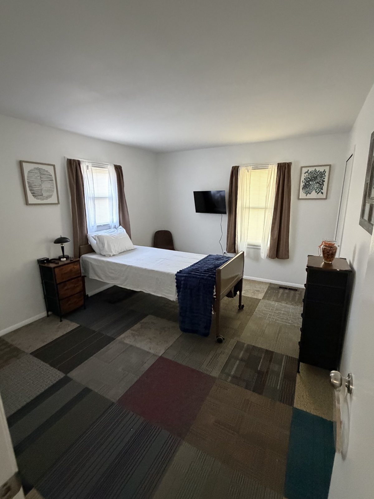 Private resident room with a bed, dresser, television and two sunlit windows